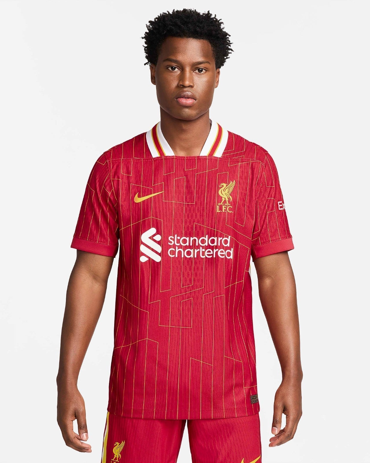 rn-image_picker_lib_temp_98520876-ccf1-4341-90cc-937fcbc9be8c | AE JERSEY Young man modeling a red Liverpool FC home jersey with yellow crest and sponsor logo, standing against a light background.