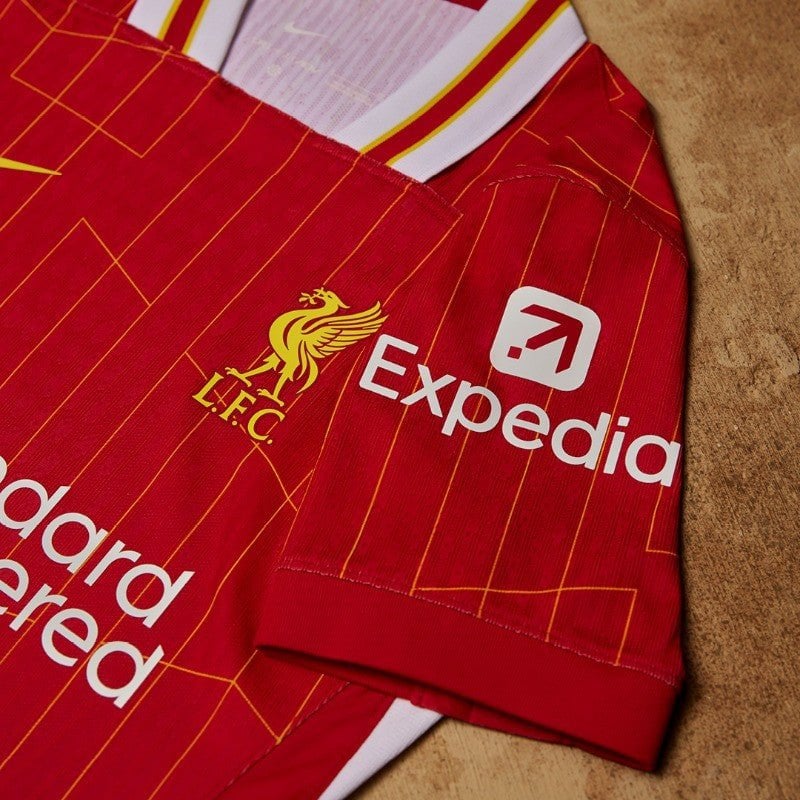 rn-image_picker_lib_temp_b07b87c9-befe-435b-8749-534c498a647b | AE JERSEY Red Liverpool FC jersey with yellow crest and Expedia sponsor logo on a wooden surface, showing team branding and design lines.