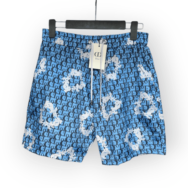 Blue shorts featuring a repeating Dior monogram pattern with white paint splatter details, displayed on a hanger with a tag attached