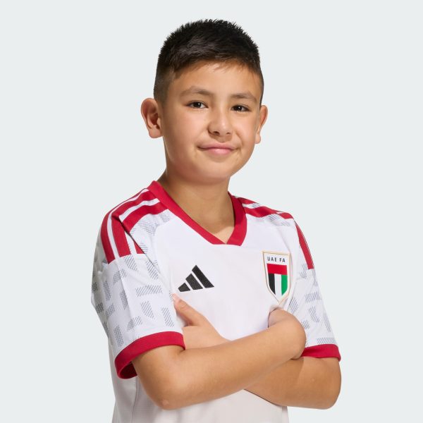 Portrait of a young boy wearing a white and red UAE FA jersey, arms crossed, facing the camera against a light gray background.