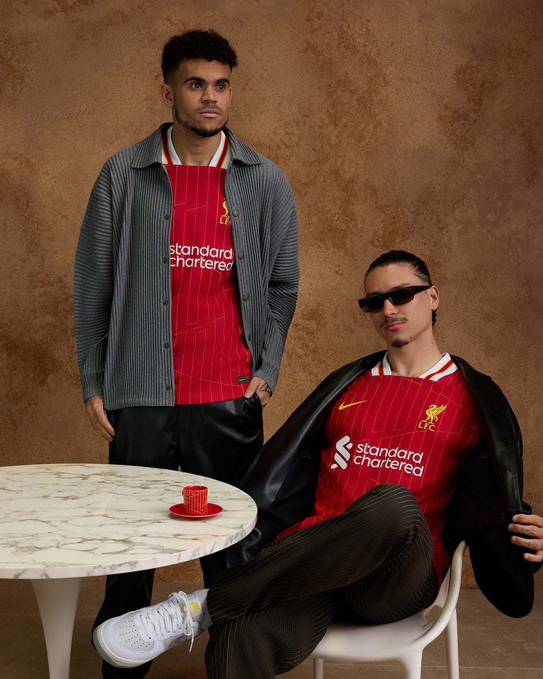 rn-image_picker_lib_temp_d463e155-5f3b-4650-8311-591d32cf4ea8 | AE JERSEY Two men in red Liverpool FC jerseys pose against a brown textured wall; one stands in a gray cardigan, the other sits in a chair wearing sunglasses and a black jacket over a jersey.