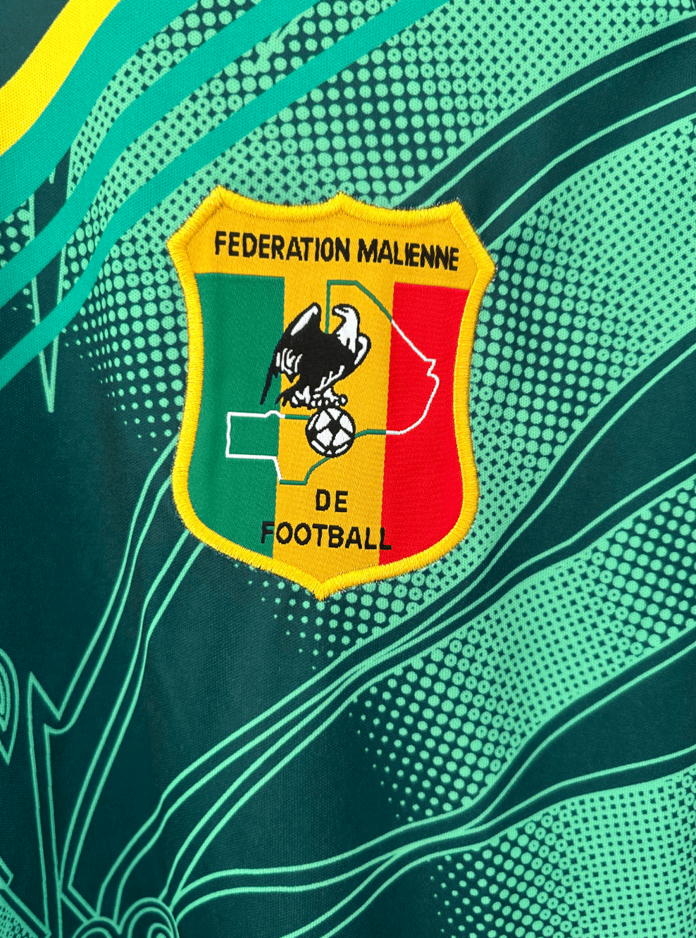 rn-image_picker_lib_temp_d75b65a8-eba5-4fcc-a042-507e32141922 | AE JERSEY Embroidered shield logo of the Fédération Malienne de Football: green, yellow, red vertical stripes, black eagle with a football, on a teal fabric background.