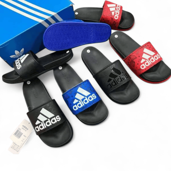 Assorted Adidas slide sandals in black, blue, and red with white logos, arranged near a blue shoe box.