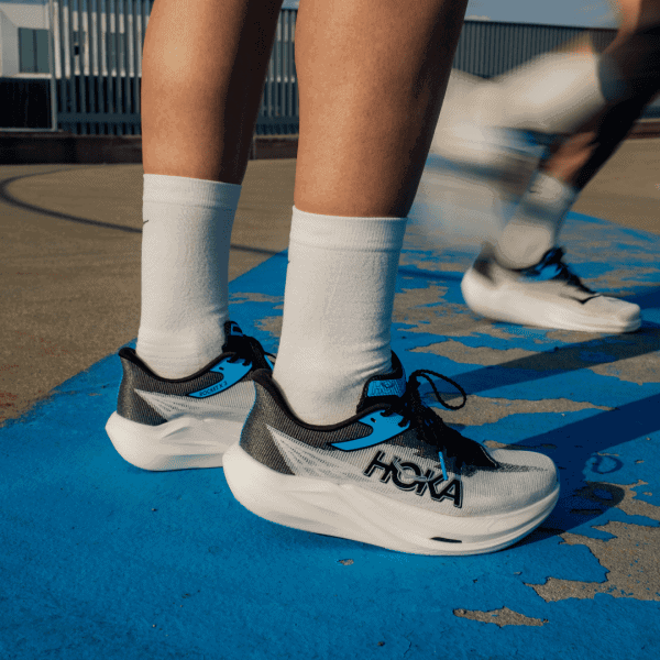 Close-up of grey and blue Hoka running shoes on a blue track, worn with white socks and bare calves visible on either side.