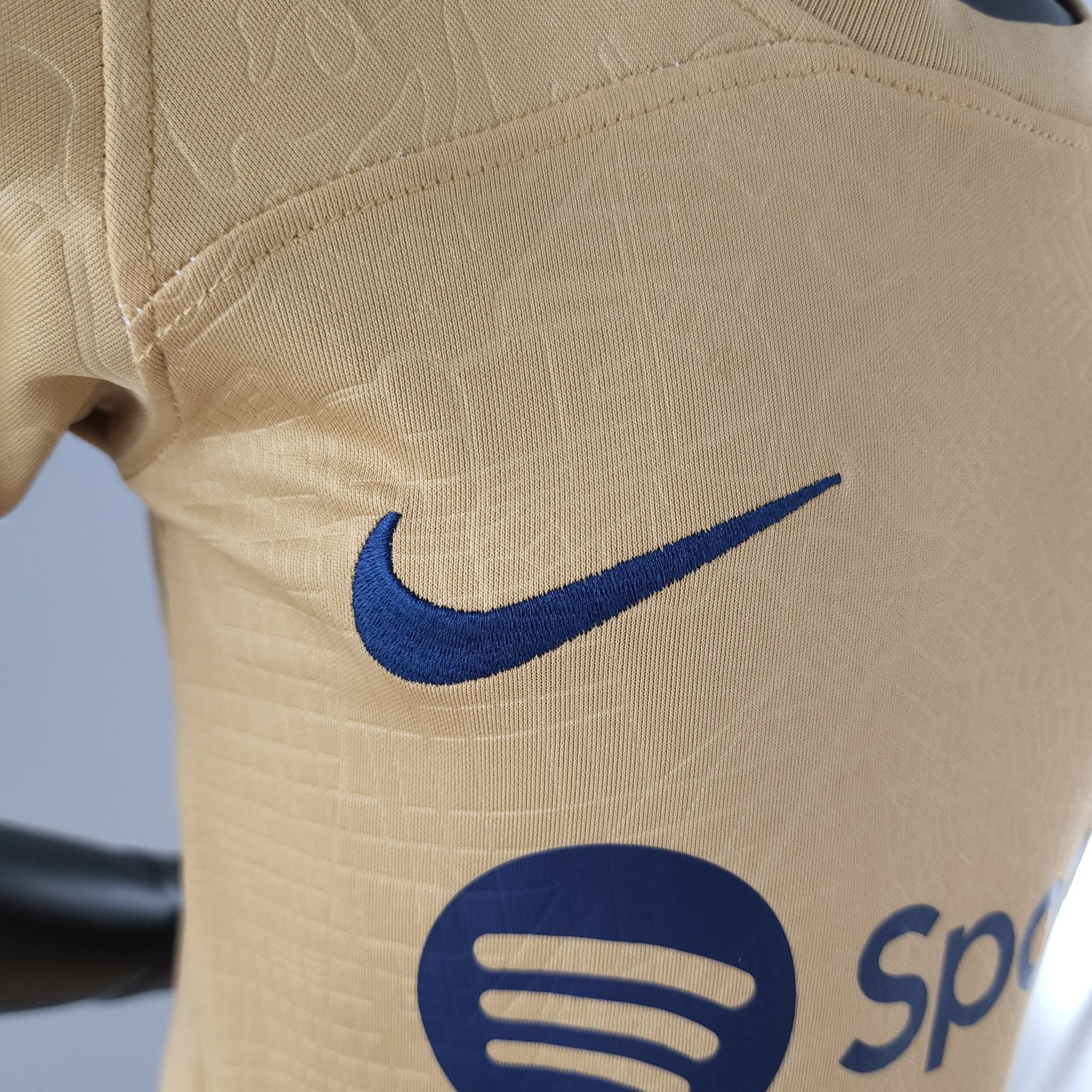 rn-image_picker_lib_temp_f5a35daa-f6b8-478b-9363-27afa9facafc | AE JERSEY Close-up of beige fabric showing a blue Nike swoosh logo partially visible and stitching detail.
