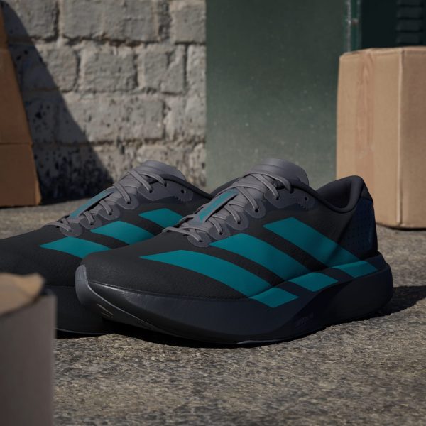 Close-up of gray running shoes with teal stripes resting on rough outdoor ground with a brick wall and cardboard boxes in the background