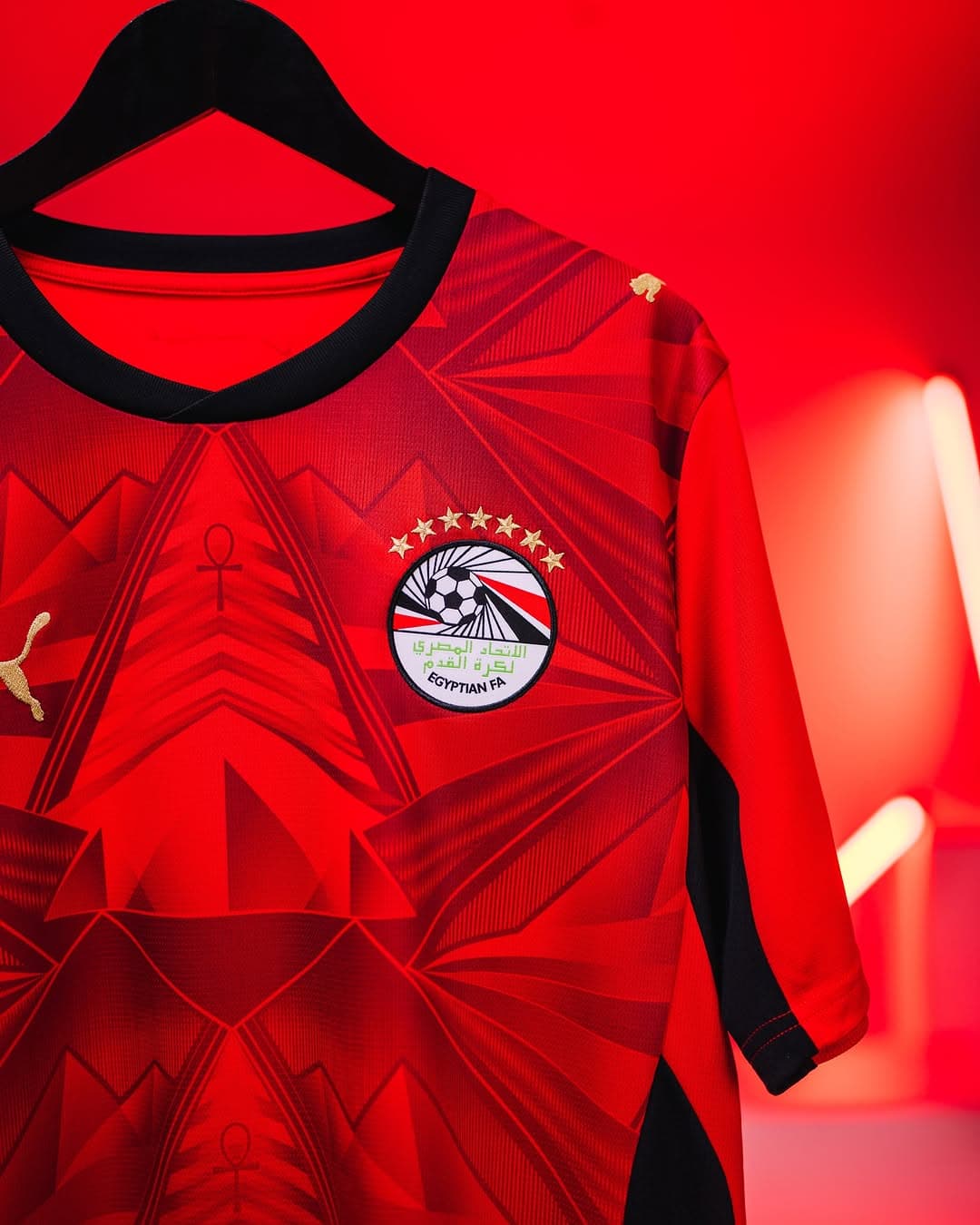 rn-image_picker_lib_temp_f8184854-e637-4c0c-8669-bad850c2cbeb | AE JERSEY Red football jersey on a black hanger, displaying the Egyptian FA crest with stars on a bold red background.