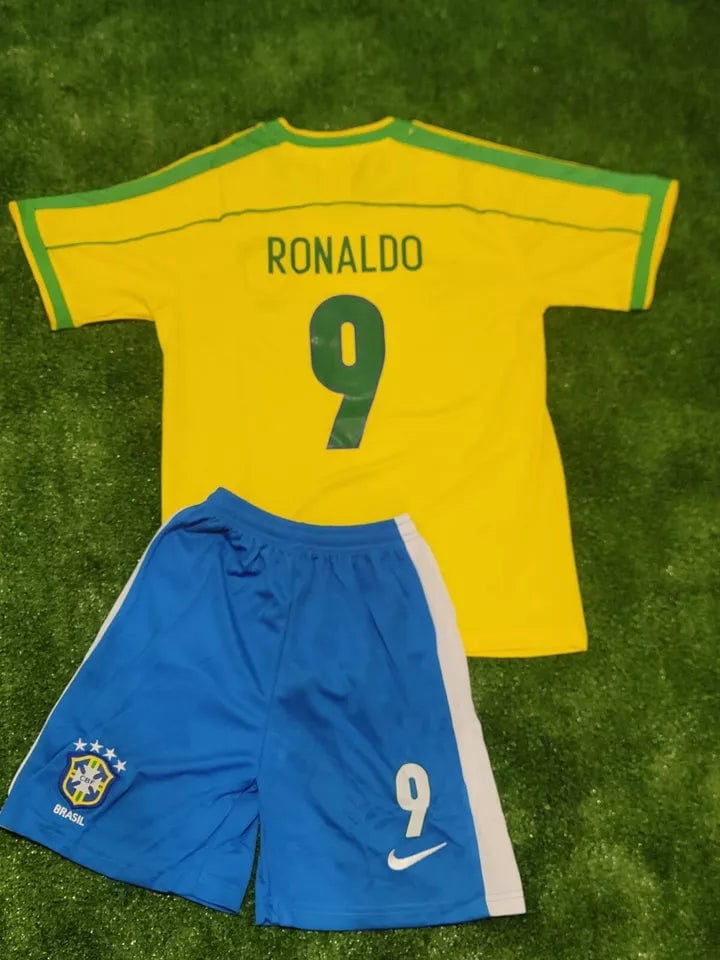 s-l960_11 | AE JERSEY Yellow Brazil soccer jersey with 'RONALDO' and number 9 laid over blue shorts on grass, celebrating a football kit set up.