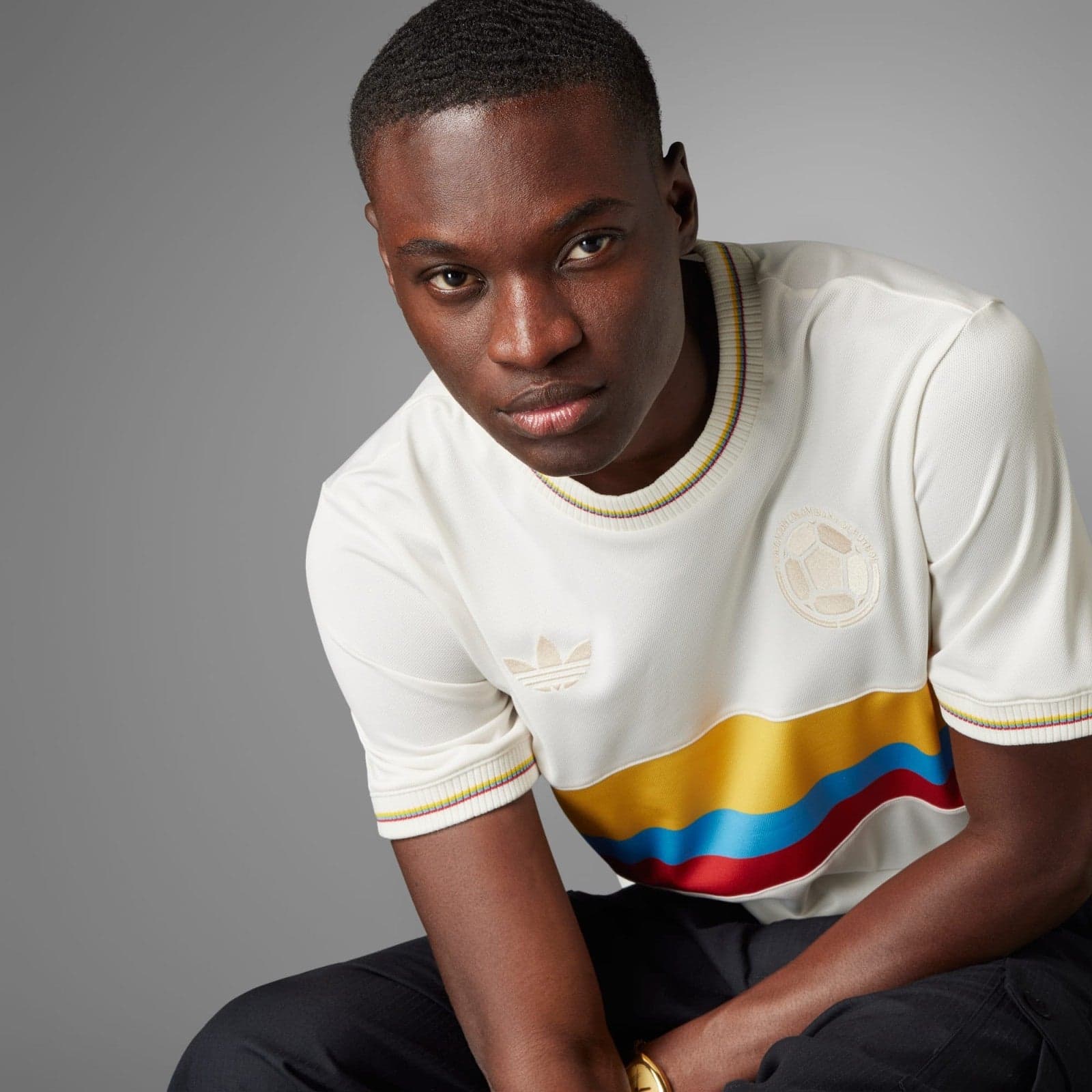 wjN7ALLYGSeg9p1 | AE JERSEY Portrait of a young man in a white sweater with rainbow stripes, seated against a gray backdrop and looking slightly to the side.